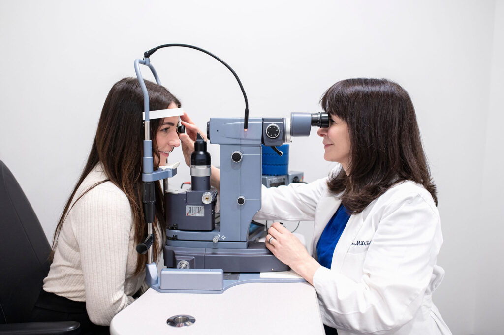 Eye doctor conducting a comprehensive eye exam using a slit lamp microscope while a female patient rests her chin on the exam device in a clinic.