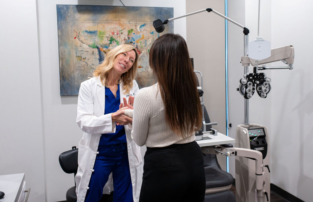 Eye doctor speaking with a female patient during a consultation in an exam room, holding the patient’s hands while discussing vision care.